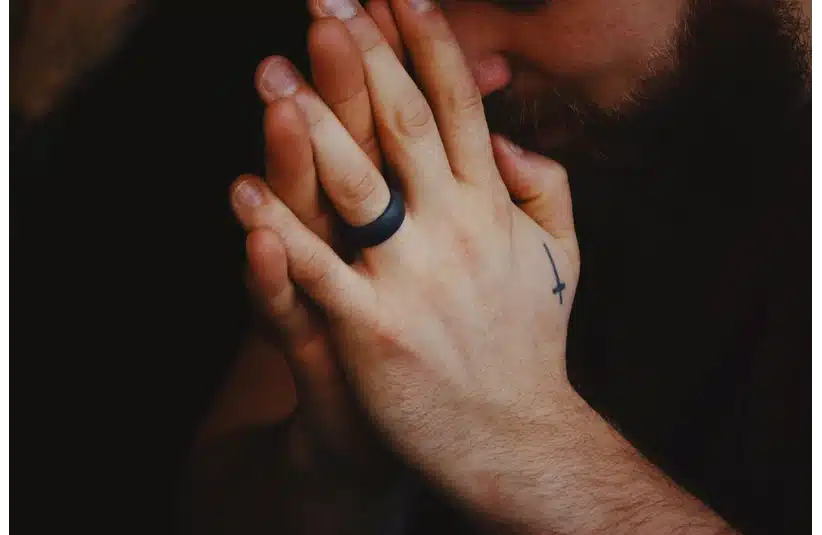 A person with a beard holding their hands in a prayer position, displaying a small cross tattoo and a black ring, conveying a contemplative mood.,