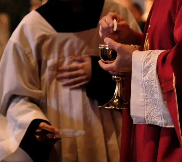 A priest in red robes holds a chalice and Eucharist wafer during a religious ceremony, assisted by an altar server in white attire, creating a solemn atmosphere. Lent, Easter, Christian, Catholic, church, mass, holy communion