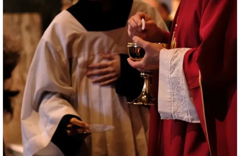A priest in red robes holds a chalice and Eucharist wafer during a religious ceremony, assisted by an altar server in white attire, creating a solemn atmosphere. Lent, Easter, Christian, Catholic, church, mass, holy communion