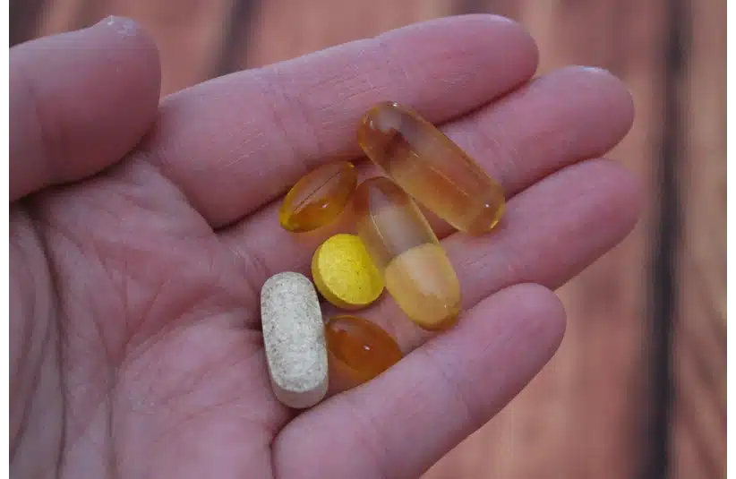 A hand holds various vitamins, including transparent softgels, a round yellow tablet, and a beige oblong pill, against a blurred wooden background.