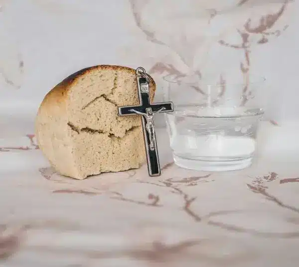A piece of bread, a small cross, and a glass of water sit on a patterned cloth. The scene conveys simplicity and spirituality, Lent. Fat Tuesday is the day before Lent begins.