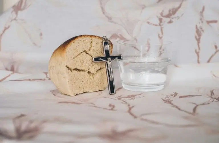A piece of bread, a small cross, and a glass of water sit on a patterned cloth. The scene conveys simplicity and spirituality, Lent. Fat Tuesday is the day before Lent begins.