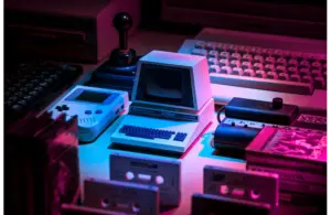 A retro setup with a small vintage computer, Game Boy, joystick, keyboard, and cassette tapes. Neon pink and blue lighting creates a nostalgic vibe. Black innovators who revolutionized gaming.
