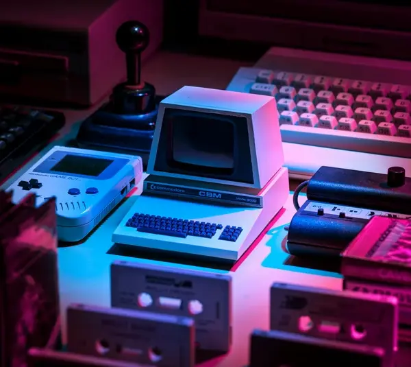 A retro setup with a small vintage computer, Game Boy, joystick, keyboard, and cassette tapes. Neon pink and blue lighting creates a nostalgic vibe. Black innovators who revolutionized gaming.