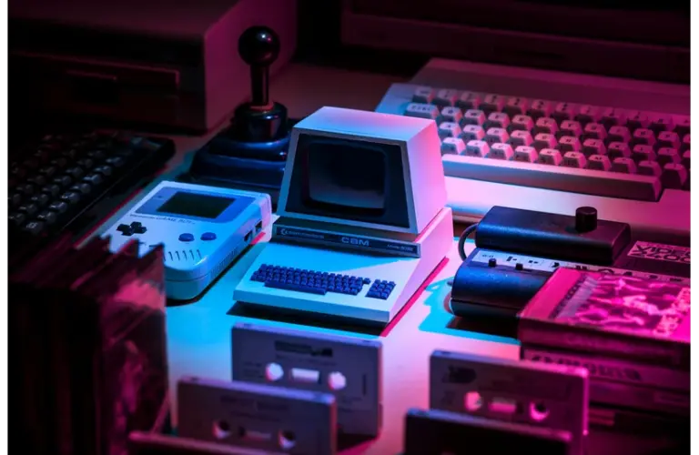 A retro setup with a small vintage computer, Game Boy, joystick, keyboard, and cassette tapes. Neon pink and blue lighting creates a nostalgic vibe. Black innovators who revolutionized gaming.