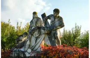 A white stone statue depicting a biblical scene of three figures supporting a cross, surrounded by greenery and vibrant red bushes under a clear sky. Ash Wednesday, Catholic, Christian, Jesus, crucifixion