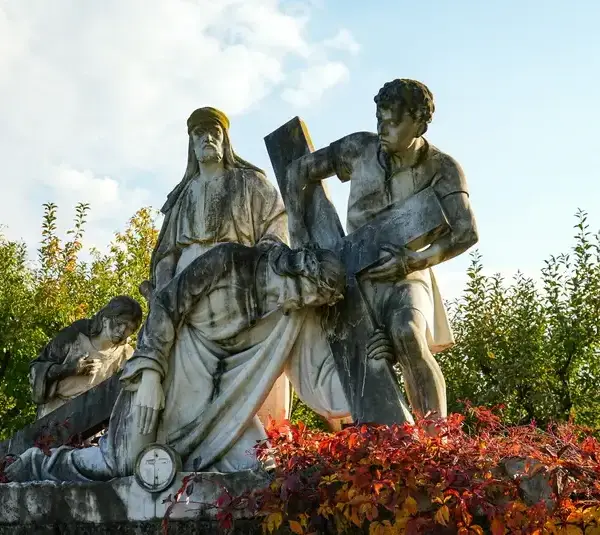 A white stone statue depicting a biblical scene of three figures supporting a cross, surrounded by greenery and vibrant red bushes under a clear sky. Ash Wednesday, Catholic, Christian, Jesus, crucifixion