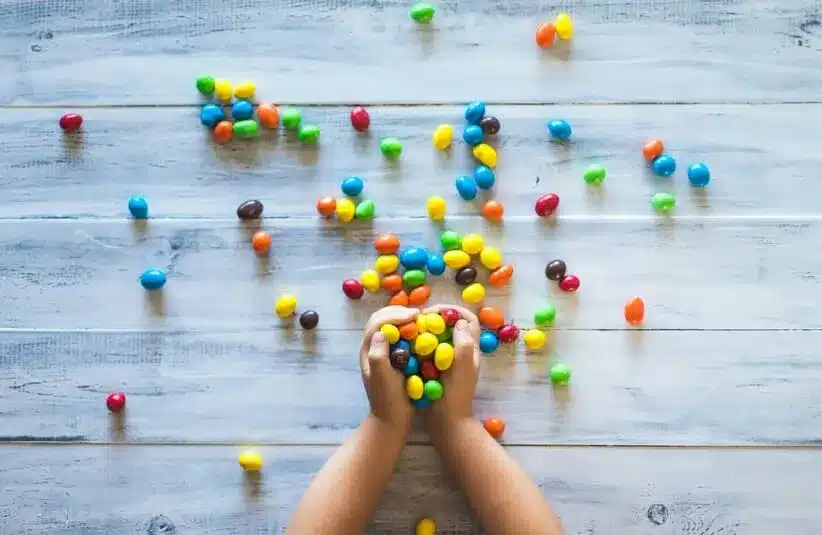 image of Child's hands holding colorful candies M&M's scattered on a light wooden table. The vibrant candy colors create a playful and joyful atmosphere.
