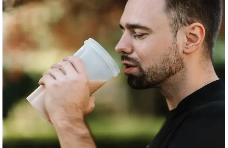A man with a beard enjoys a protein shake from a white cup, outdoors. The background is blurred with earthy tones, creating a calm and relaxed mood. best supplements for men