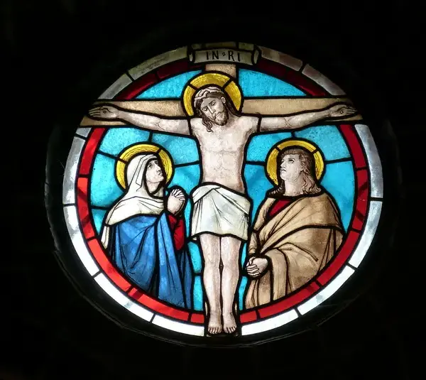 Stained glass window depicting a crucifixion scene. Central figure on a cross with halo, flanked by two figures in robes. Blue and red hues convey solemnity. Lent, Christian, Catholic, church, Jesus