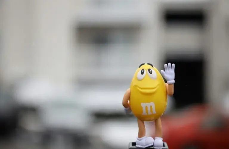 image of a small yellow M&M's figurine stands with a friendly smile, waving. It features cartoon-like eyes and limbs, set against a blurred urban background.