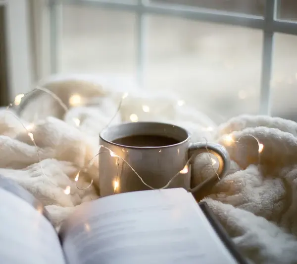 a cozy setting with a book, soft blanket, fairy lights, and a warm cup of coffee