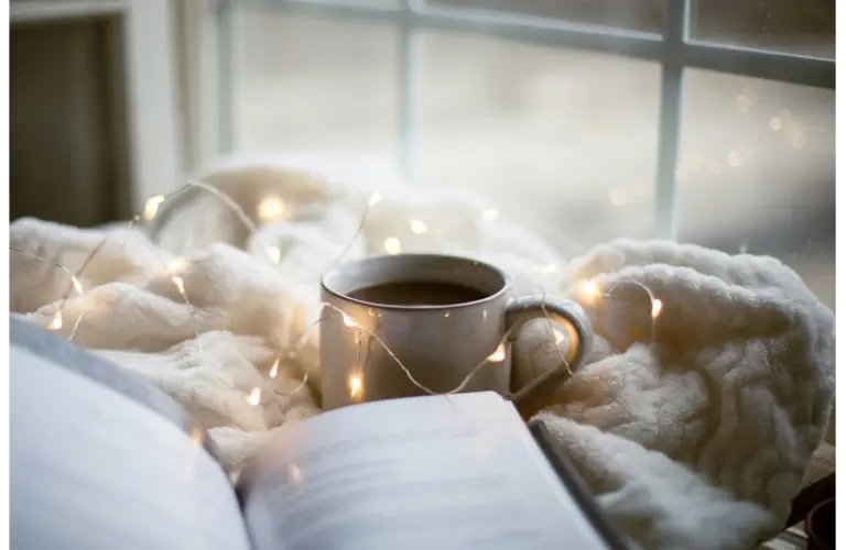 a cozy setting with a book, soft blanket, fairy lights, and a warm cup of coffee