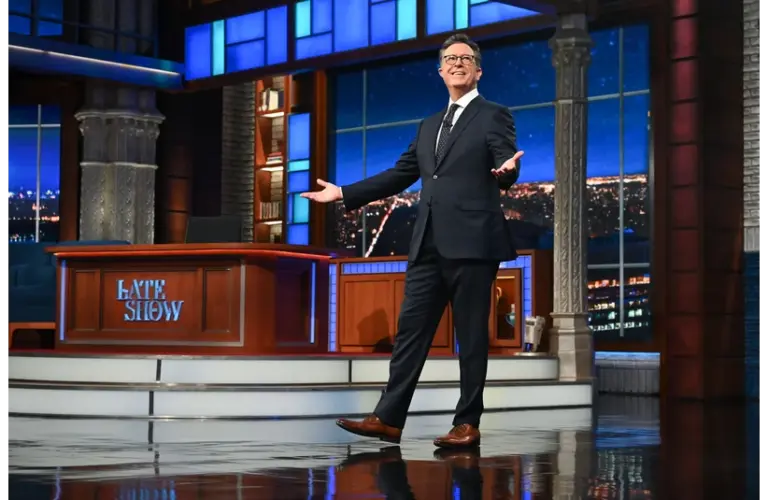 Stephen Colbert, host of The Late Show and writer for the next Lord of the Rings film, stands smiling on a brightly lit talk show stage with a cityscape backdrop. He gestures invitingly, conveying a welcoming and cheerful atmosphere.
