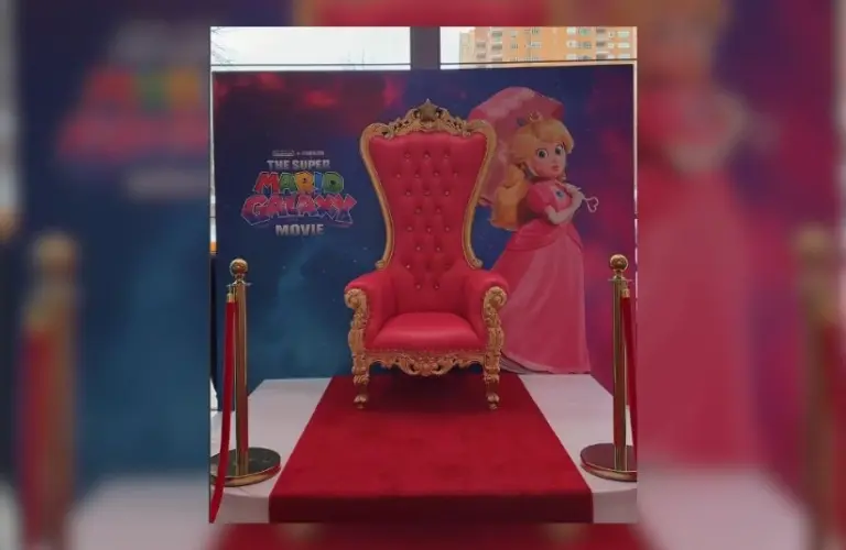 A pink plush high-back throne chair on a red carpet in front of a Super Mario Galaxy advertisement that has Princess Peach on it