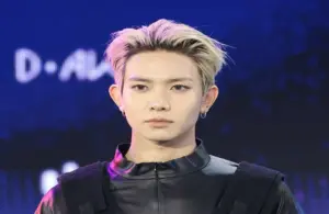 ENHYPEN Heeseung with frosted blonde hair and dark roots. He is wearig a dark shirt with a black vest on top.