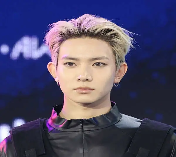 ENHYPEN Heeseung with frosted blonde hair and dark roots. He is wearig a dark shirt with a black vest on top.