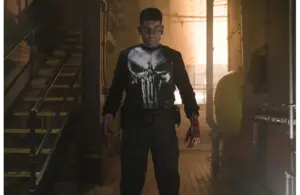 Frank Castle, aka The Punisher, played by Jon Bernthal, stands in a dimly lit industrial setting, wearing a shirt with a large white skull emblem. His expression is intense, and his hands are stained, suggesting a recent struggle. The atmosphere is tense and gritty.