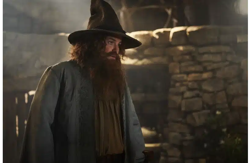 Rory Kinnea as Tom Bombadil, dressed as a wizard with a long beard and pointed hat, stands in a rustic stone-walled room. The atmosphere is mysterious and medieval.