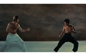 One man (Chuck Norris) in white pants on the left and one man (Bruce Lee) with black pants face each other in fighting stances