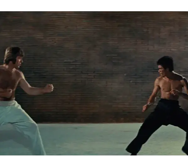 One man (Chuck Norris) in white pants on the left and one man (Bruce Lee) with black pants face each other in fighting stances