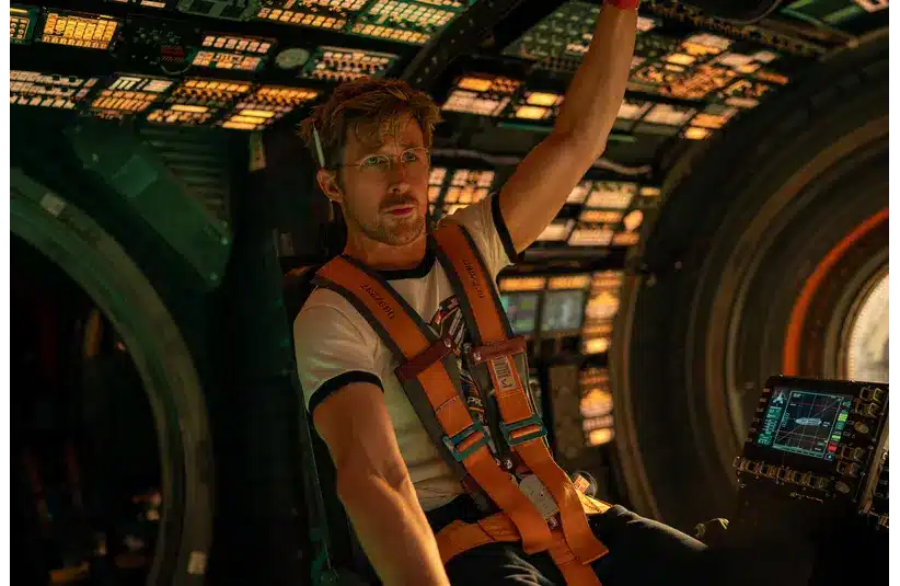 Man in a spaceship cockpit, wearing a harness and white T-shirt, reaches overhead. The control panel is lit with colorful buttons, conveying tension.