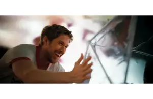 Ryan Gosling in "Project Hail Mary (2026)" smiles joyfully, reaching out towards a reflective, angular surface. The scene is brightly lit, conveying a sense of happiness and warmth.