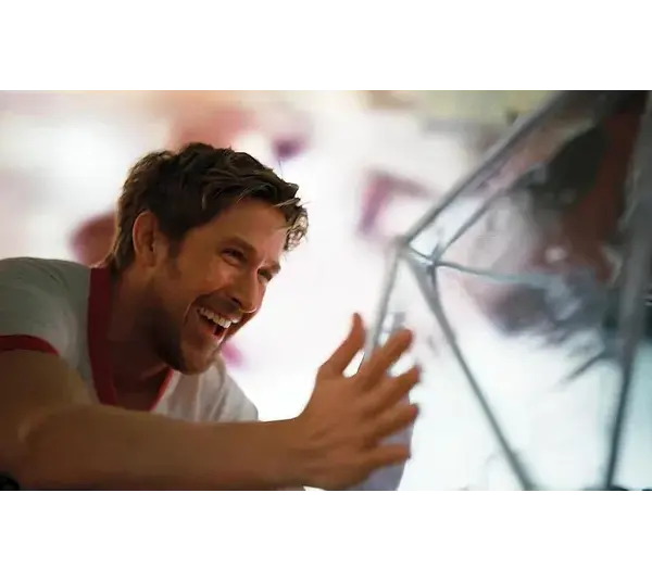 Ryan Gosling in "Project Hail Mary (2026)" smiles joyfully, reaching out towards a reflective, angular surface. The scene is brightly lit, conveying a sense of happiness and warmth.