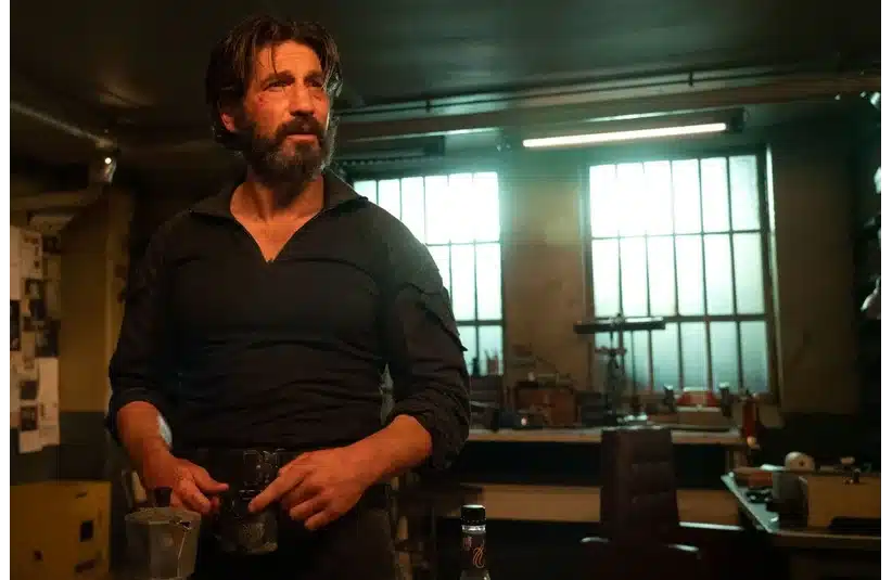 Frank Castle, aka The Punisher, played by Jon Bernthal, with a beard, stands in a dimly lit workshop, illuminated by a window. He appears contemplative, holding a cup, surrounded by tools and equipment.