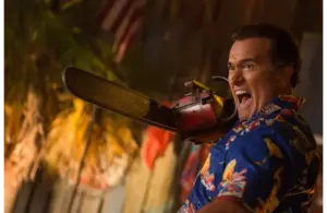 Bruce Campbell a blue patterned shirt, holds a chainsaw and shouts, with an American flag and palm leaves visible in the background.