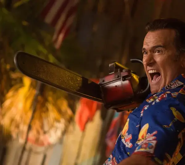Bruce Campbell a blue patterned shirt, holds a chainsaw and shouts, with an American flag and palm leaves visible in the background.