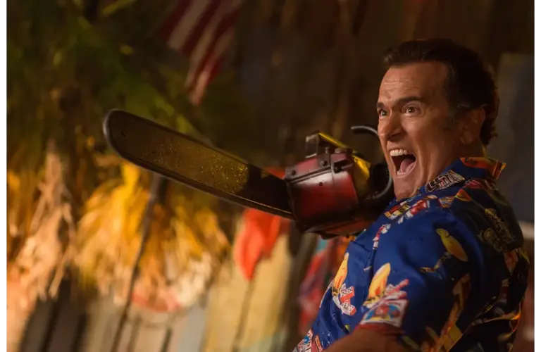 Bruce Campbell a blue patterned shirt, holds a chainsaw and shouts, with an American flag and palm leaves visible in the background.