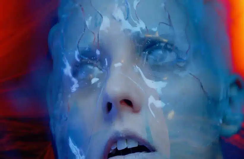 A close-up of a person's face covered in a translucent blue material, eyes wide, conveying anxiety, unseeing. Red hues blend with blue, adding a surreal atmosphere.
