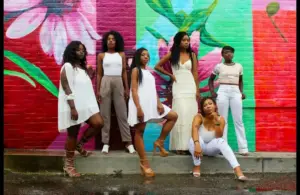 Six women pose confidently in front of a vibrant, floral mural. Their stylish white outfits contrast against the colorful red and green background. Women's History Month
