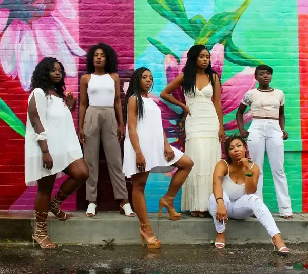 Six women pose confidently in front of a vibrant, floral mural. Their stylish white outfits contrast against the colorful red and green background. Women's History Month