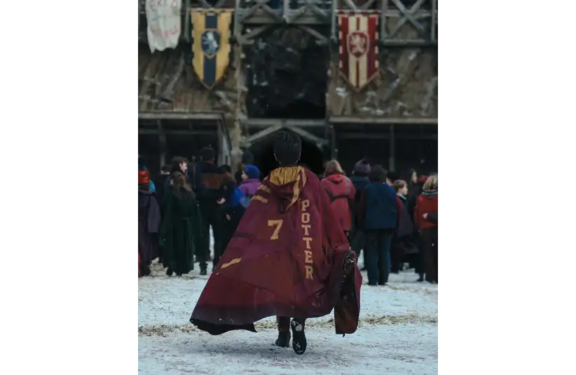 Dominic McLaughlin as Harry Potter in a red cloak with "Potter" and "7" walks across a snowy field, heading towards a rustic building with two banners. Crowd in the background.