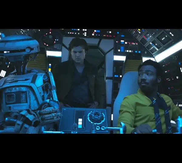 A humanoid robot with Donald Glover and Alden Ehrenreich are in a futuristic spaceship cockpit filled with glowing controls. They appear focused and determined.