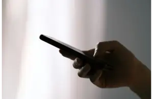 A hand holds a smartphone against a softly lit, blurred background. The image conveys a sense of focus and connectivity in a subdued, calm setting. text scam, fake recall, text message, smartphone