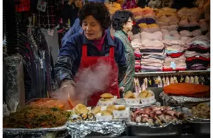 A woman wearing a red apron cooks at a bustling market stall with steaming food, surrounded by colorful textiles, conveying a lively, busy atmosphere. Women's History Month, food traditions