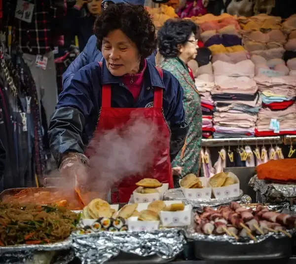 A woman wearing a red apron cooks at a bustling market stall with steaming food, surrounded by colorful textiles, conveying a lively, busy atmosphere. Women's History Month, food traditions