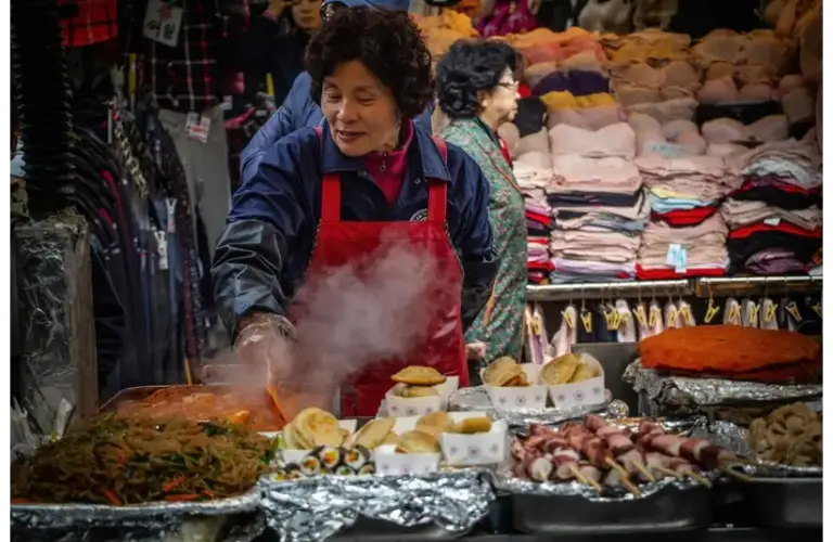 A woman wearing a red apron cooks at a bustling market stall with steaming food, surrounded by colorful textiles, conveying a lively, busy atmosphere. Women's History Month, food traditions