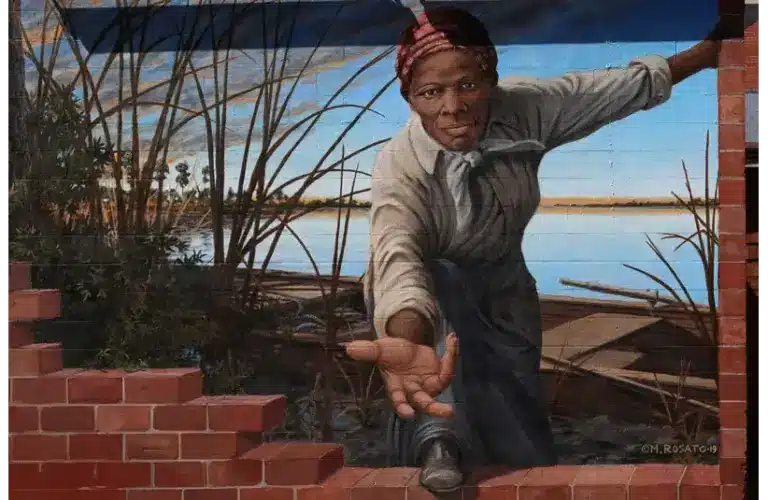 Mural of Harriet Tubman reaching out through a brick wall, set against a riverbank with reeds. The expression is determined and inviting, suggesting courage and guidance. Women's History Month