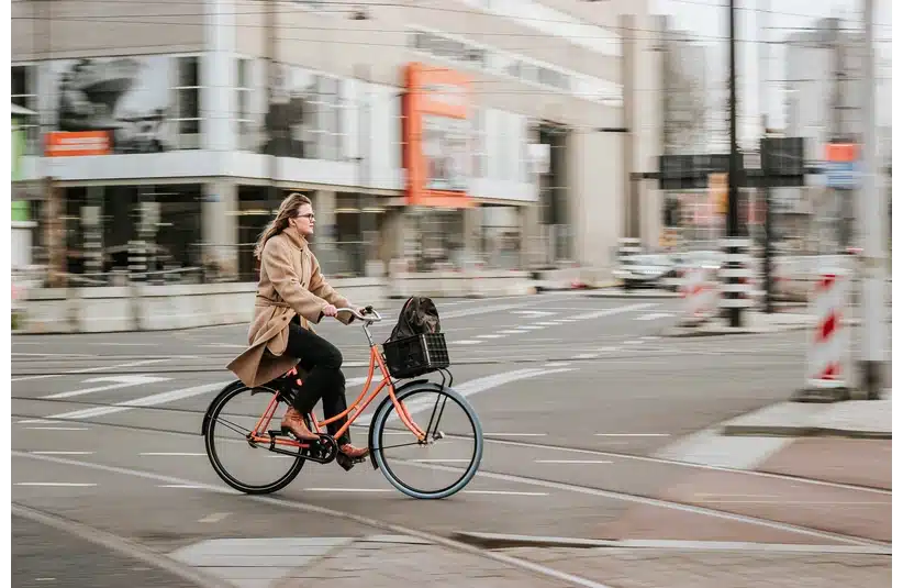 A person in a beige coat rides a bright orange bicycle through a city street, with a blurred background conveying motion and urban life.