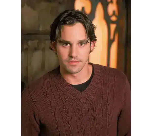 Young man, Nicholas Brendon, with dark hair in a brown cable-knit sweater stands indoors against a rustic background. He has an attentive and calm expression.