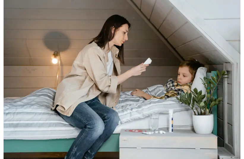 A concerned woman sits beside a sick child in bed, holding a thermometer. A bedside table holds a glass, medicine, and a plant, under soft lighting.