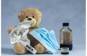 A plush teddy bear holds tissues, a face mask, and a thermometer, accompanied by a medicine bottle and a cup. The scene suggests a theme of caring for illness., recall of children's ibuprofen