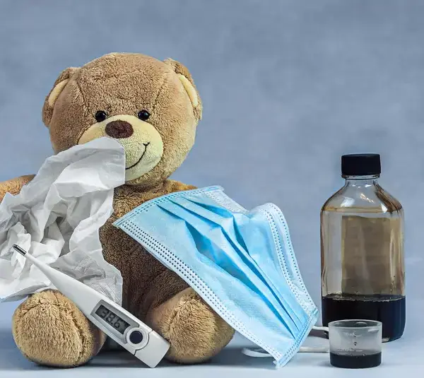 A plush teddy bear holds tissues, a face mask, and a thermometer, accompanied by a medicine bottle and a cup. The scene suggests a theme of caring for illness., recall of children's ibuprofen