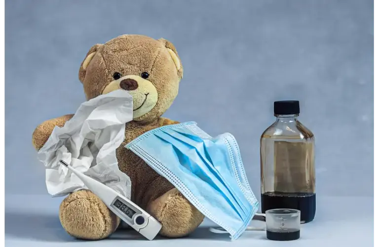A plush teddy bear holds tissues, a face mask, and a thermometer, accompanied by a medicine bottle and a cup. The scene suggests a theme of caring for illness., recall of children's ibuprofen