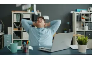 A man is celebrating success by throwing papers in the office. Lots of money falling on him in an office while sitting at a desk enjoying wealth. Happy and rich youth—a financial wellness concept.