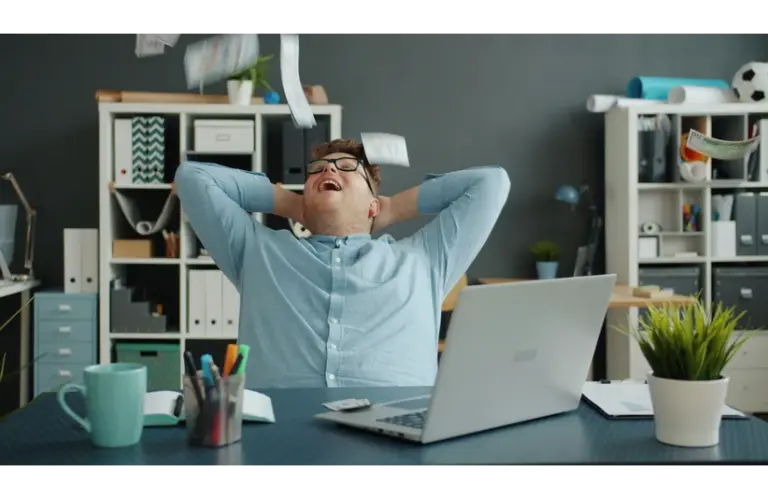 A man is celebrating success by throwing papers in the office. Lots of money falling on him in an office while sitting at a desk enjoying wealth. Happy and rich youth—a financial wellness concept.